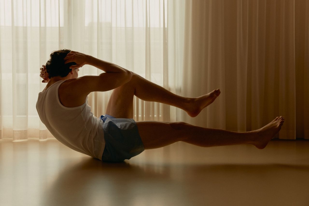 Man in a white tank top holding a Pilates curl-up on a wooden floor in front of sheer curtains